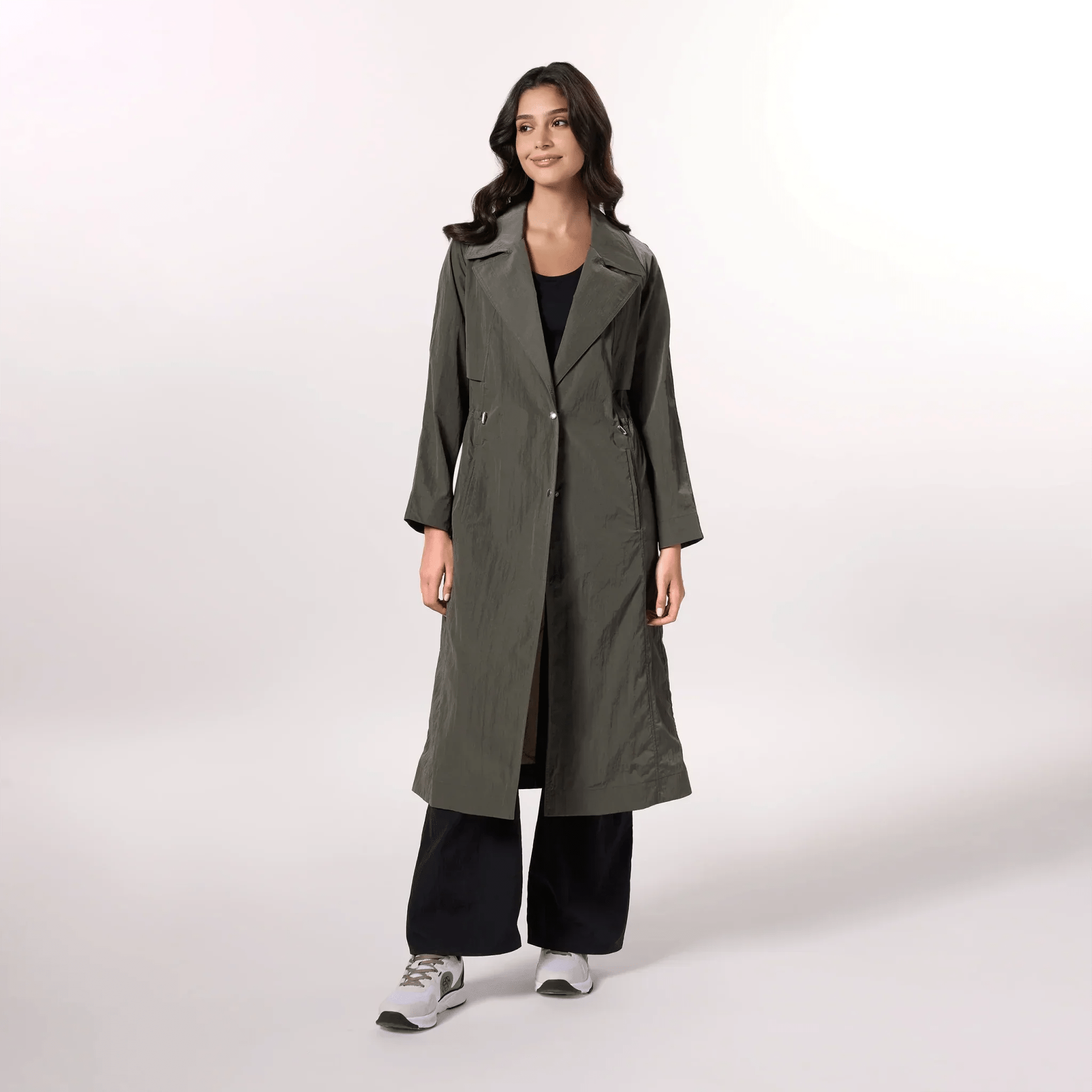 A model wearing an olive trench coat with a fluid silhouette & tie waist - عارضة أزياء ترتدي معطف ترينش باللون زيتوني بقصّة انسيابية وحزام ربط على الخصر