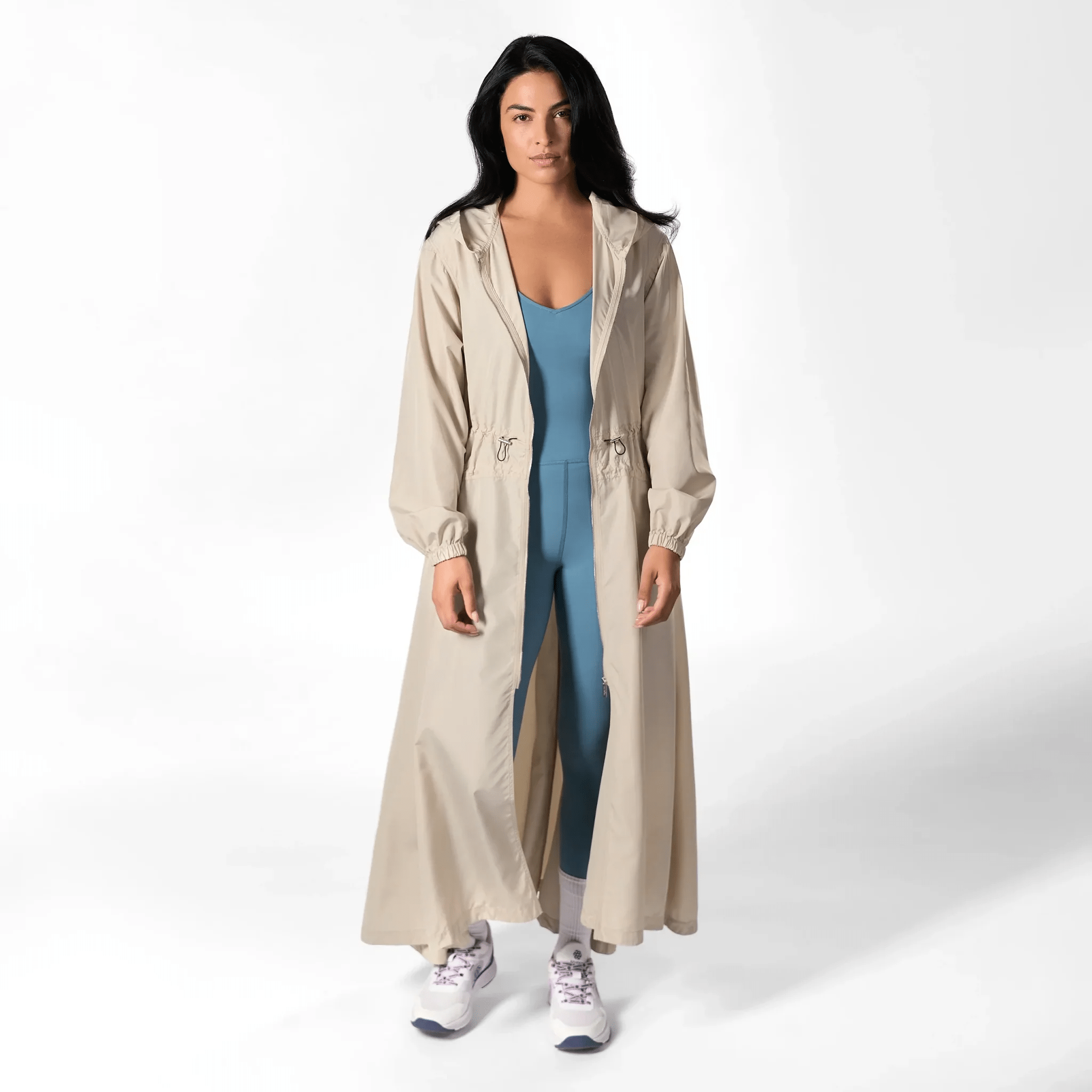 A model wearing a seasonal stone color hooded parka over a teal jumpsuit - عارضة أزياء ترتدي سترة باركا باللون الحجري بغطاء رأس فوق جمبسوت أزرق مخضر